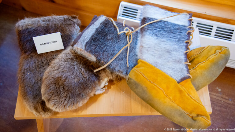 Fur mittens and yellow leather moccasin-style footwear with cord ties displayed on a museum table.