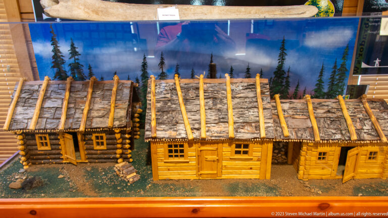 Scale model of several connected log buildings with bark roofs and small windows against a painted forest backdrop.