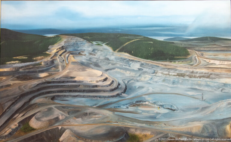 Aerial view of a vast terraced open-pit mine with winding haul roads, surrounded by forest and distant water.