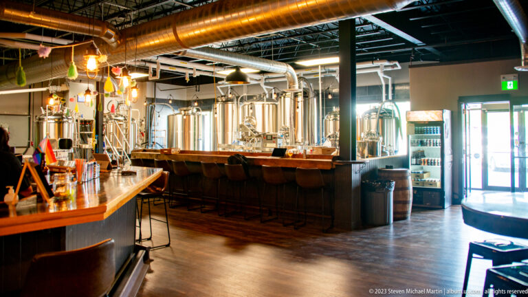 Craft brewery taproom with a long bar, stools, and stainless-steel brewing tanks beneath exposed ductwork.