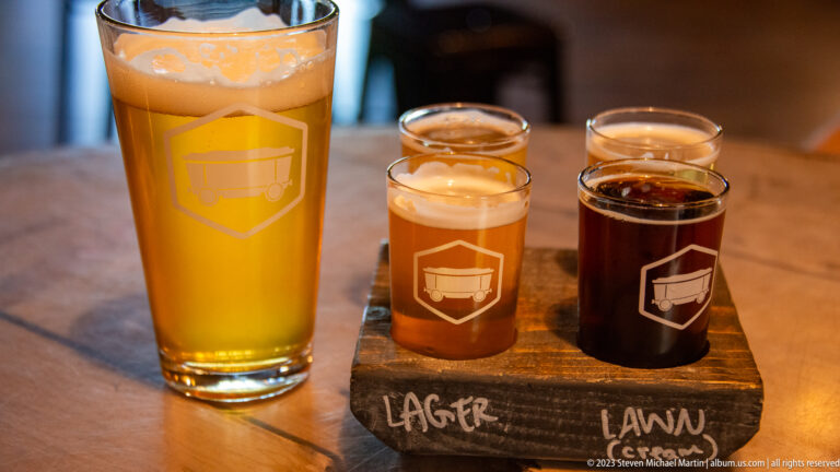 Golden pint beside four small beer samples ranging from amber to dark brown on a wooden tasting board.
