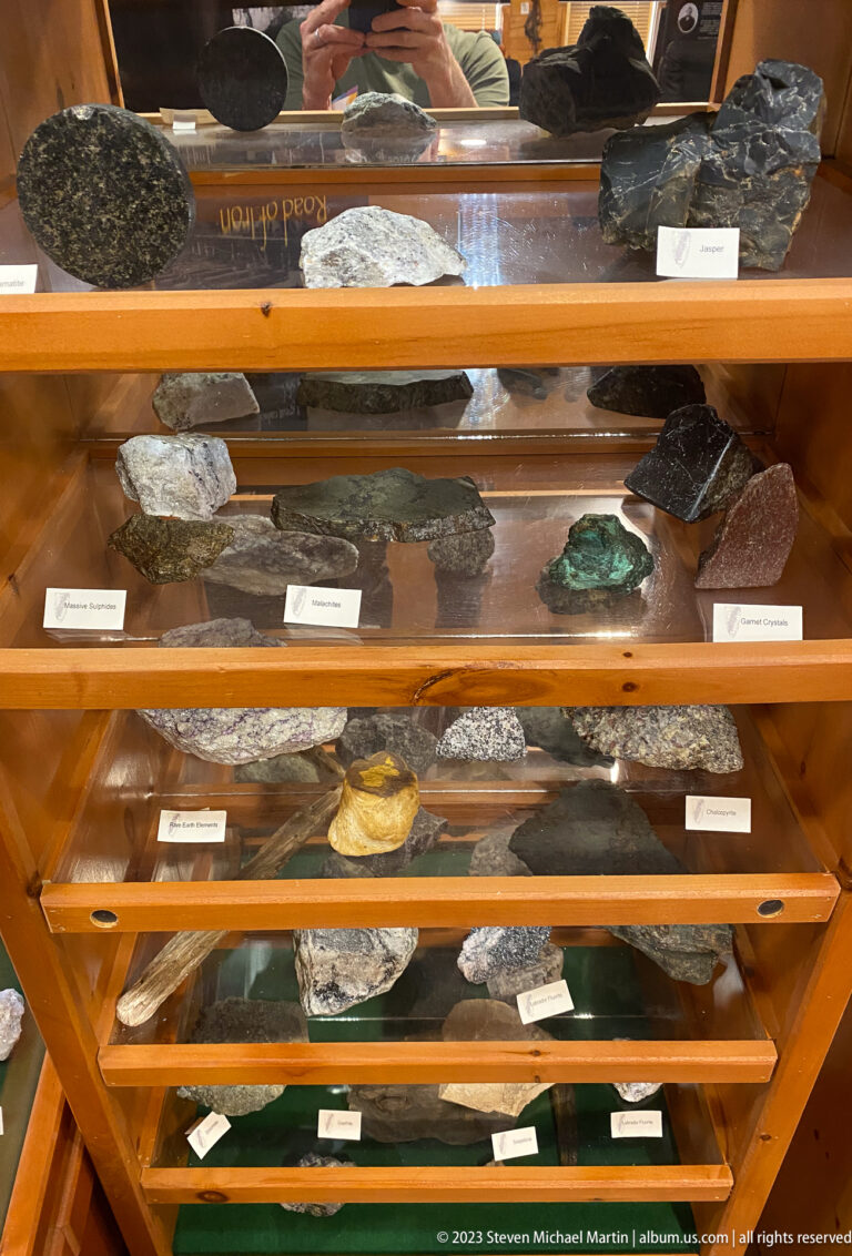 Wooden display case filled with labeled rock and mineral specimens arranged across several glass shelves.