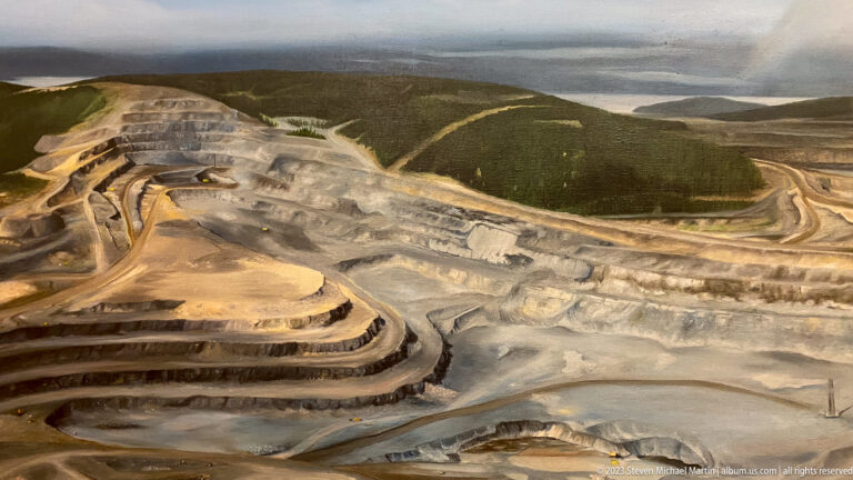 Elevated view of a vast terraced open-pit mine bordered by forest and distant lakes.