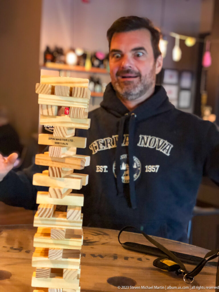 Smiling man behind a tall, slightly leaning tower of wooden stacking blocks on a tabletop.