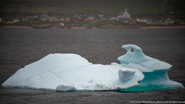 Large sculpted iceberg with a raised crest and turquoise undercut edge, village shoreline in the distance.