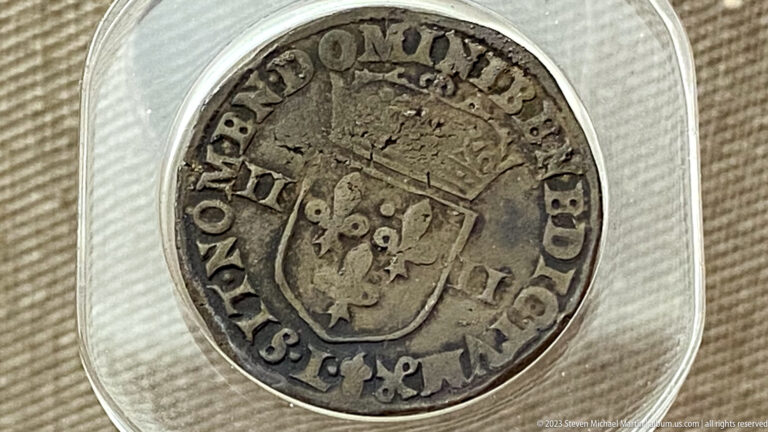 Silver-colored coin with a crowned shield and worn circular lettering displayed in a clear holder.