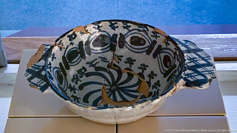 Reconstructed cream ceramic bowl with two side handles and dark green, black, and reddish painted decoration.