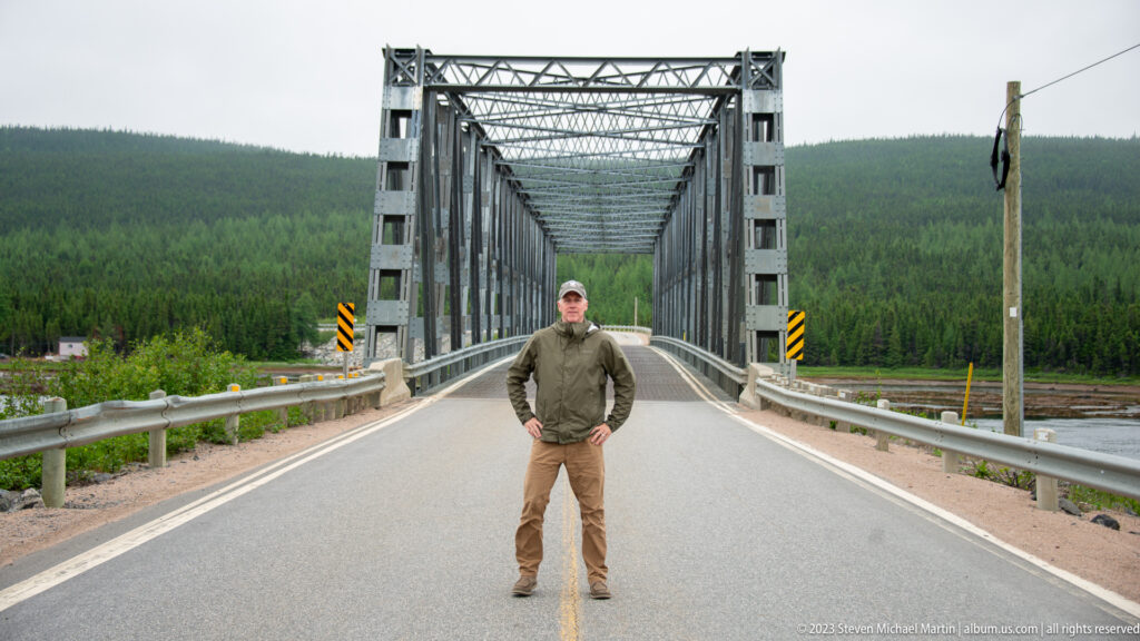 Traveler standing on the centerline at the entrance to a gray steel through-truss bridge with forested hills behind.