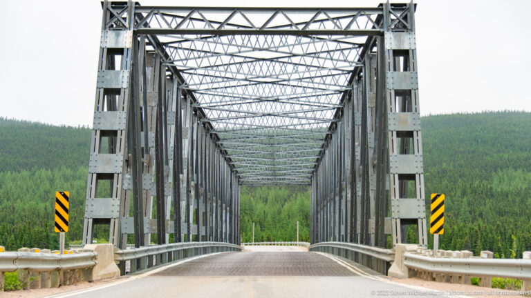 Empty gray steel through-truss bridge viewed straight on, with repeating overhead bracing and forest beyond.