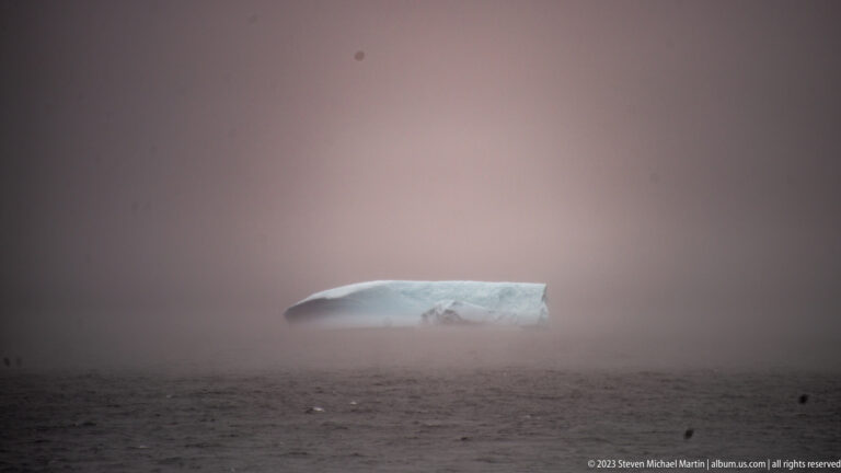 Small pale iceberg barely visible through dense fog above dark, textured seawater.