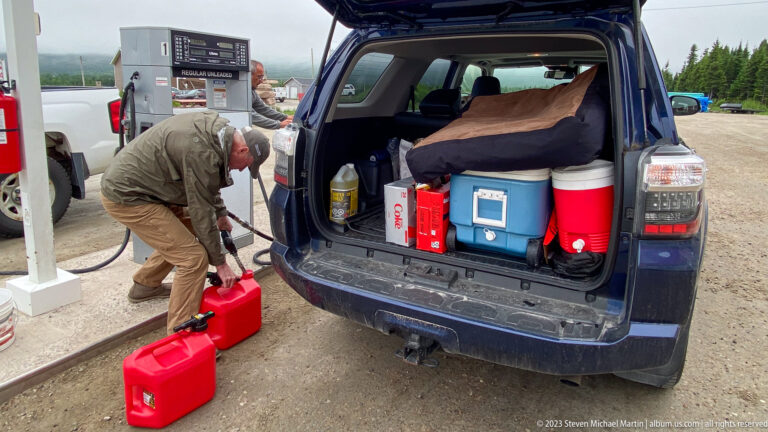 Traveler handling a red fuel can beside an open SUV packed with coolers, supplies, and additional fuel containers.