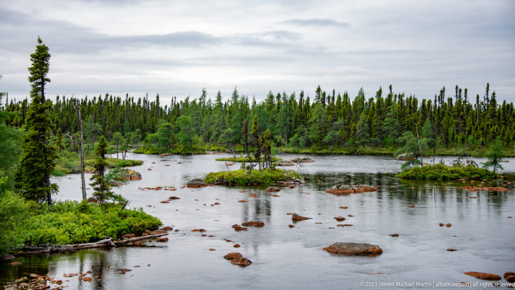 Wide shallow pond complex with reddish rocks, low islands, scattered spruce, and dense conifer forest.