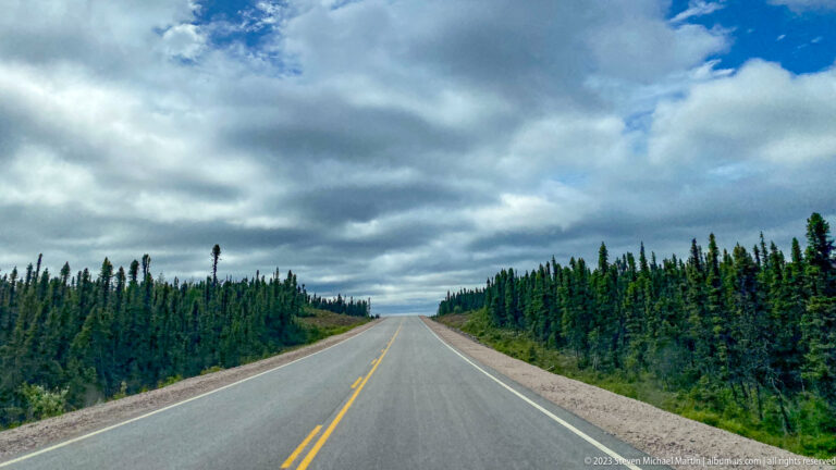 Straight two-lane highway through dense conifer forest under dramatic layered gray clouds.