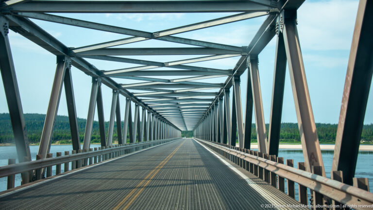View down the roadway inside a long steel through-truss bridge with water visible on both sides.