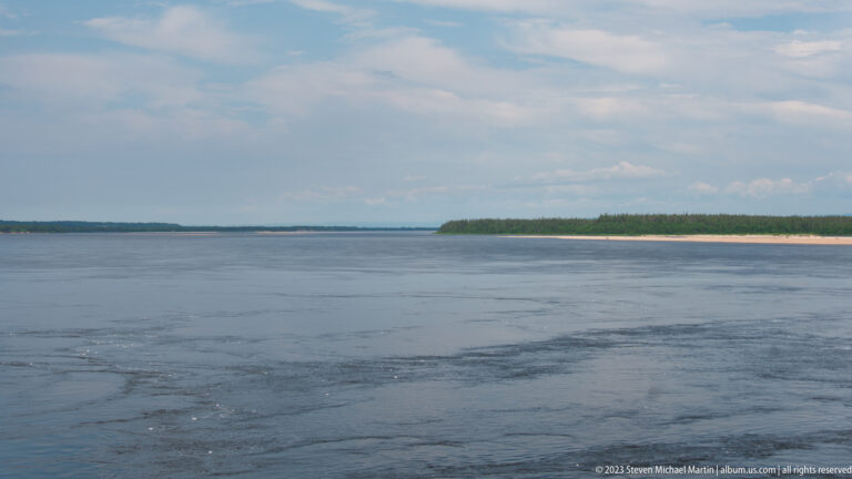 Broad blue-gray water with visible currents between low forested shores under a pale sky.