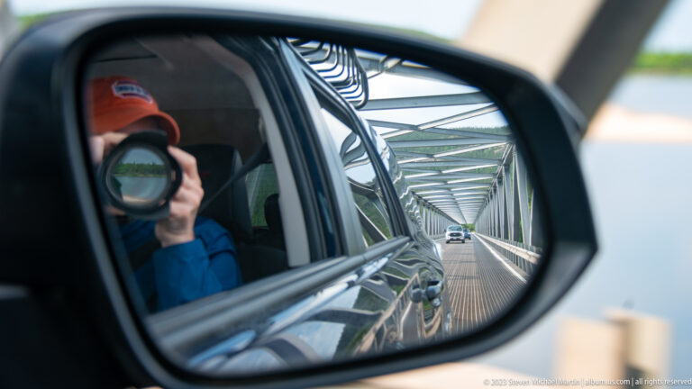 Side mirror reflecting a photographer with camera and the steel truss bridge receding behind the vehicle.