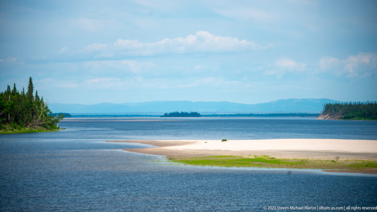 Curving pale sandbar with green vegetation in a broad blue waterway, forested shores, and hazy hills beyond.