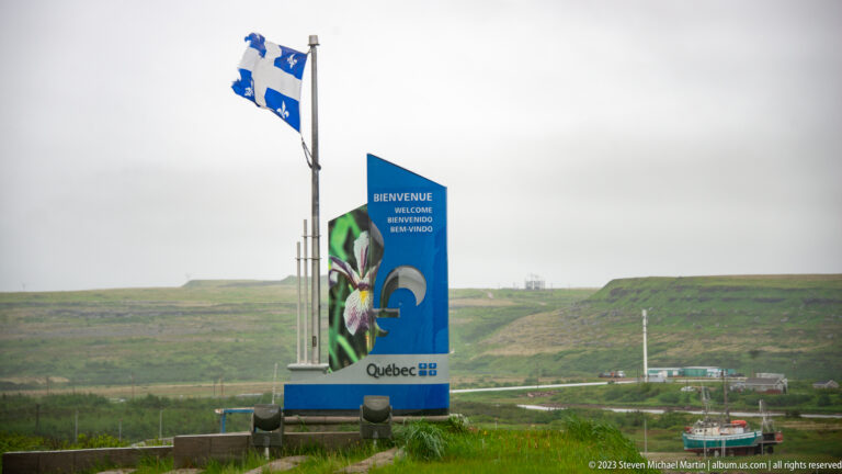 Québec flag and tall bilingual provincial welcome marker beside a road in open, treeless terrain.