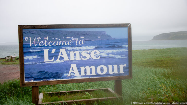 Wooden welcome sign reading 'Welcome to L’Anse Amour' beside grassy coastal terrain.