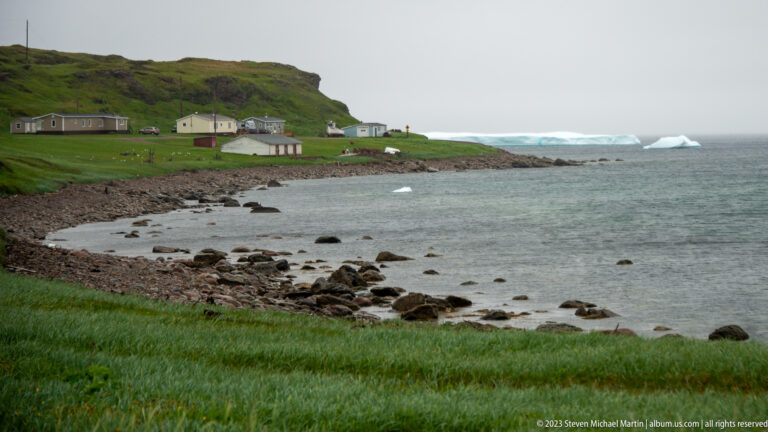 Green coastal settlement at L’Anse Amour beside a rocky shore, with several icebergs offshore.