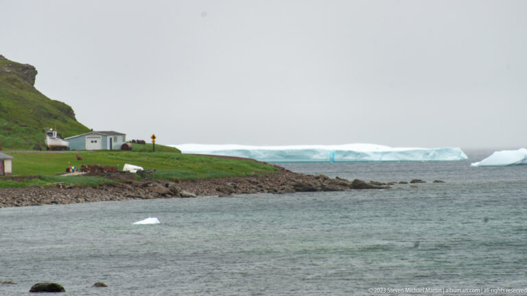 Grassy L’Anse Amour headland with small buildings at left and several broad icebergs offshore.