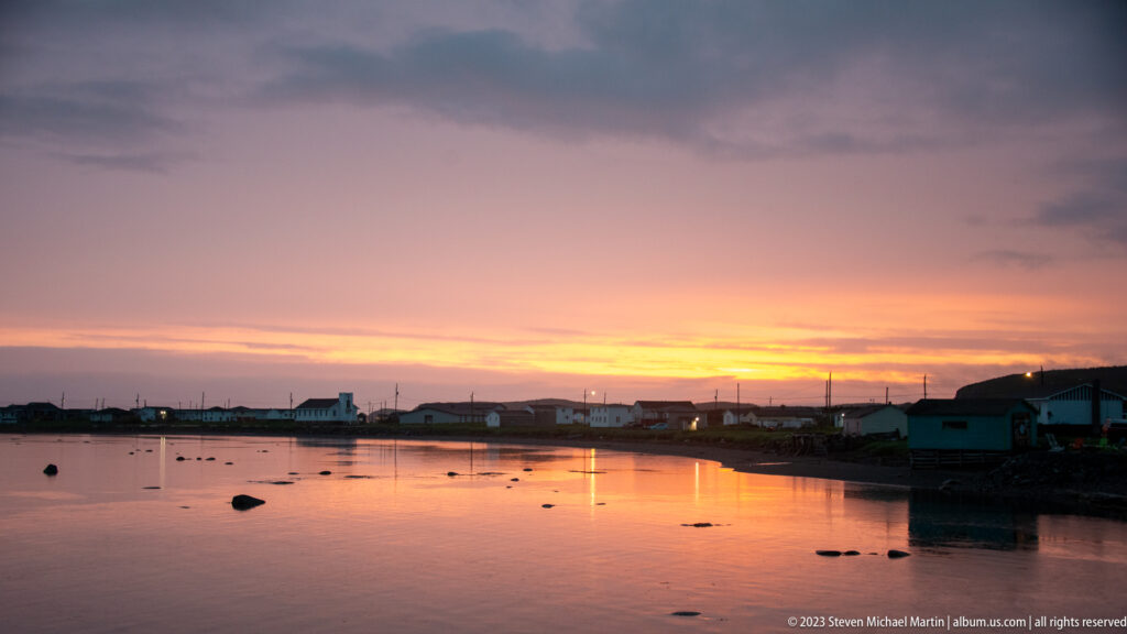 Shoreline houses and utility poles reflected in a shallow inlet under a glowing orange sunset.