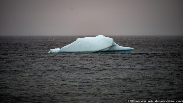 Low white iceberg with a pale blue trailing edge floating alone on dark gray water.