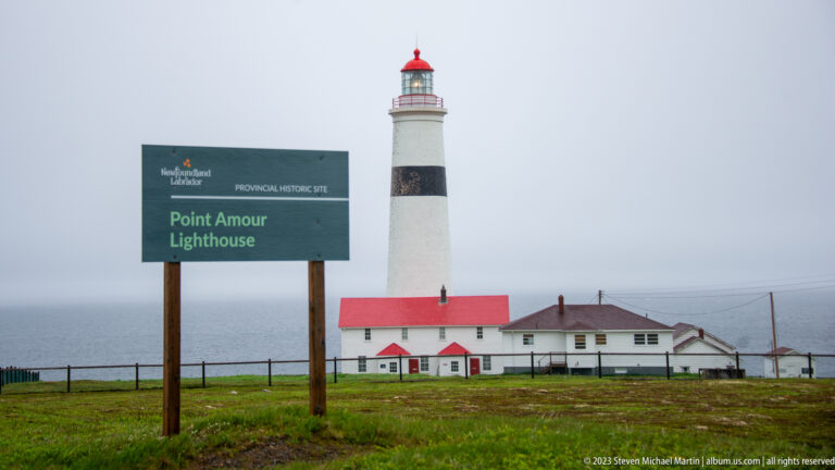 Point Amour Lighthouse sign beside a tall white tower with black band, red lantern, and red-roofed buildings.