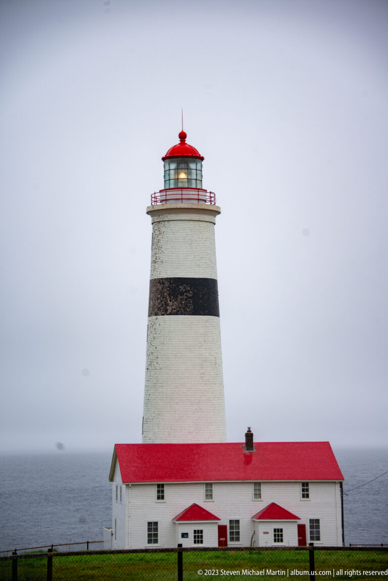 Close frontal view of Point Amour Lighthouse with white tower, black band, red lantern, and attached red-roofed dwelling.