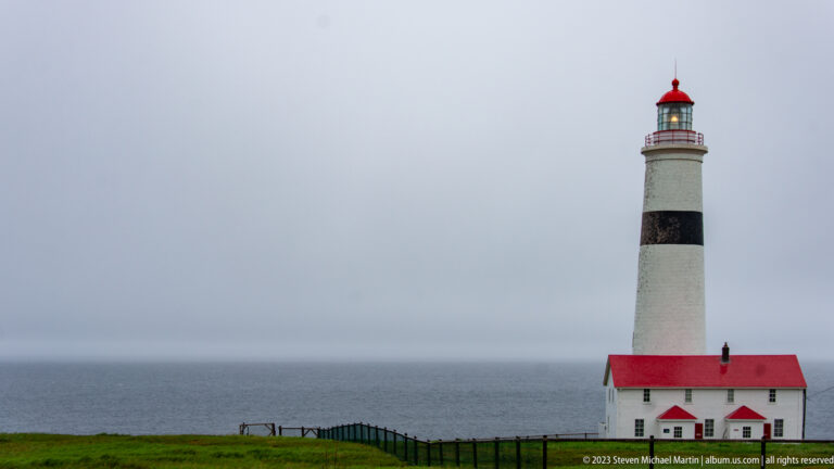 Full Point Amour Lighthouse tower rising above the sea, with black band and small red-roofed base building.