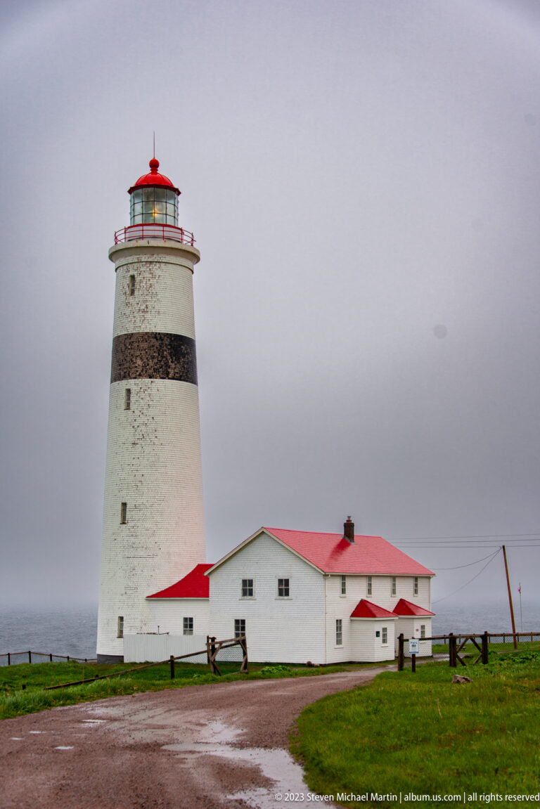 Point Amour Lighthouse and attached buildings seen from a wet gravel approach road under gray skies.