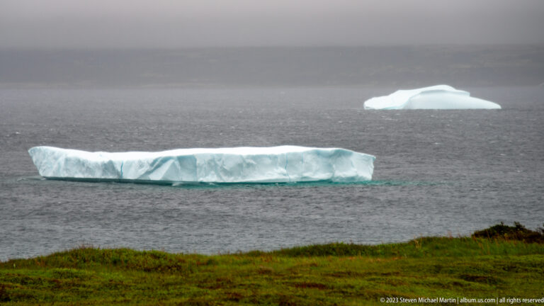 Long flat iceberg and smaller rounded iceberg offshore beyond green coastal ground under gray skies.