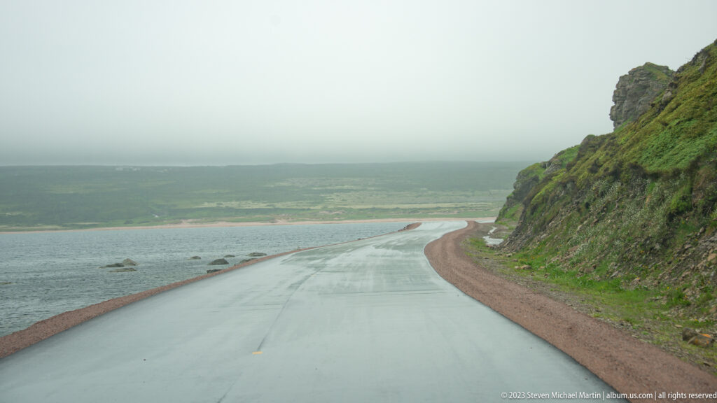 Wet coastal road curving around a steep rocky slope toward a low, misty Labrador shoreline.