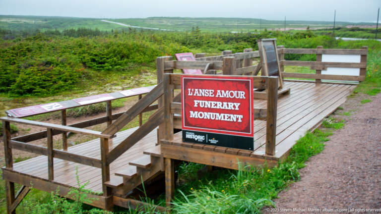 Wooden viewing platform with a red sign reading 'L’Anse Amour Funerary Monument' in a grassy archaeological site.