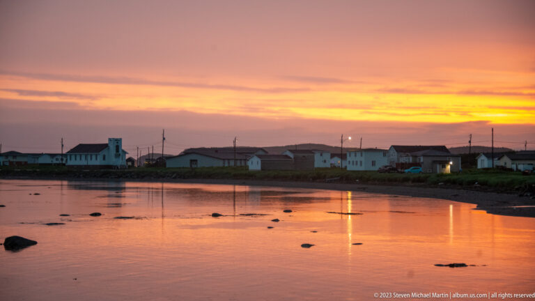 White church and low houses across a rocky tidal inlet beneath a bright orange sunset.