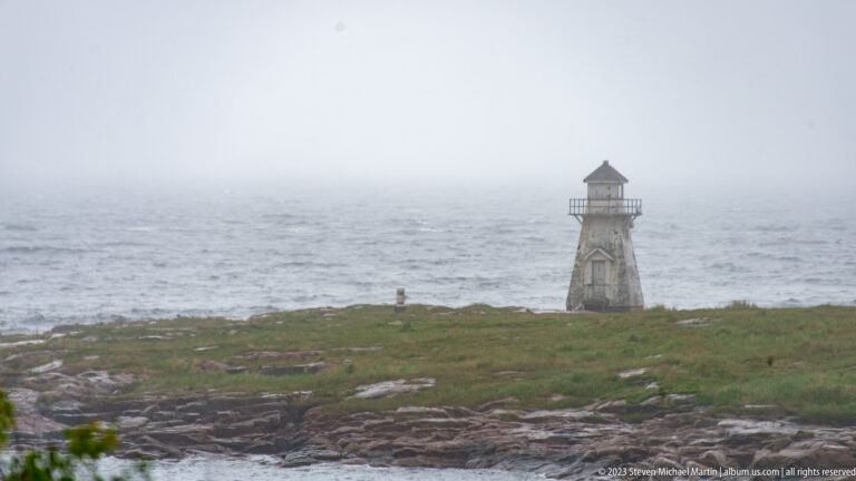 Small weathered wooden lighthouse on a low rocky point surrounded by fog and gray sea.