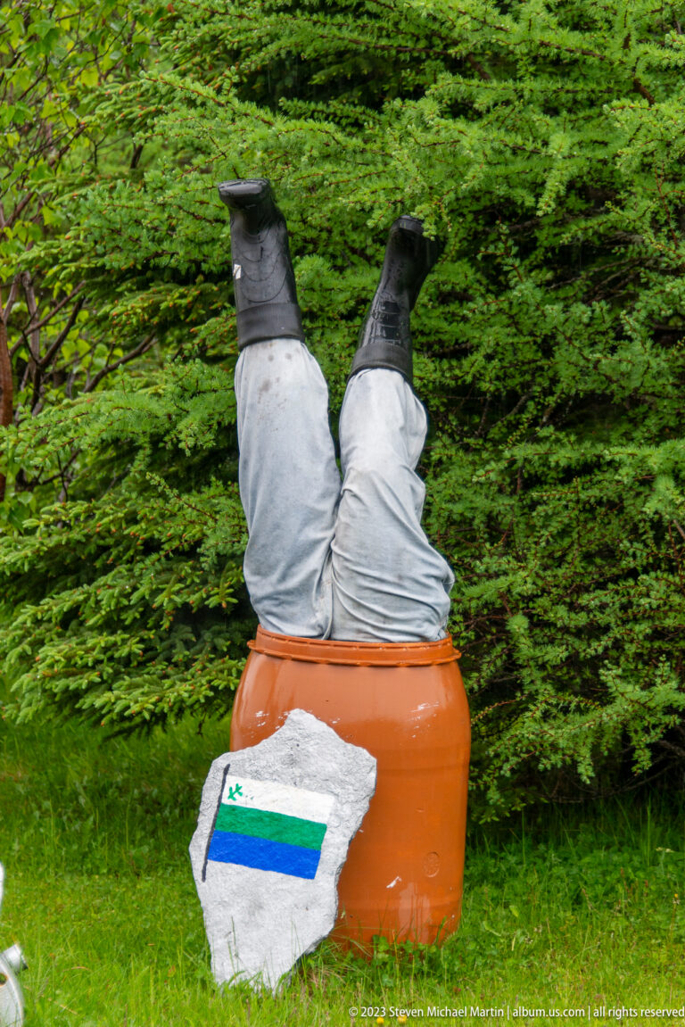 Gray trouser legs and black boots protrude upside down from an orange barrel beside a painted route marker stone.
