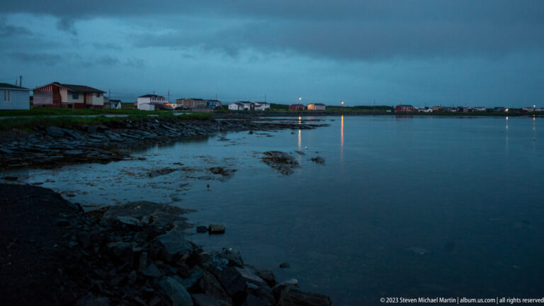 Rocky tidal shoreline at blue hour with scattered houses and distant lights reflected on calm water.