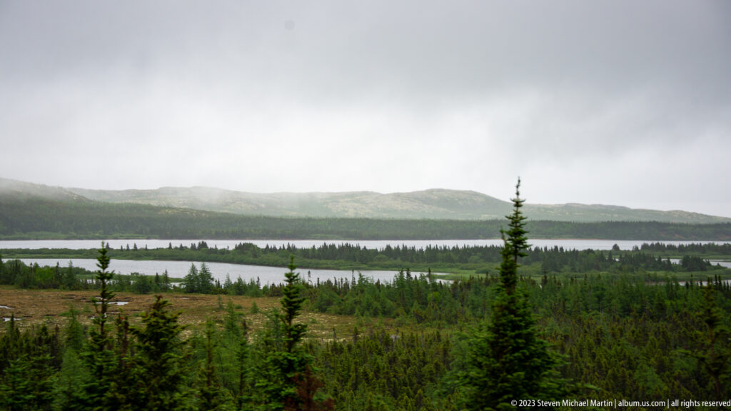 Long lake surrounded by spruce forest and low mist-covered hills under a heavy gray sky.