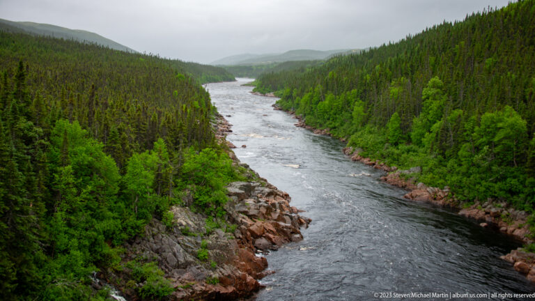 Broad dark river flowing between steep spruce-covered slopes with exposed reddish rock along both banks.