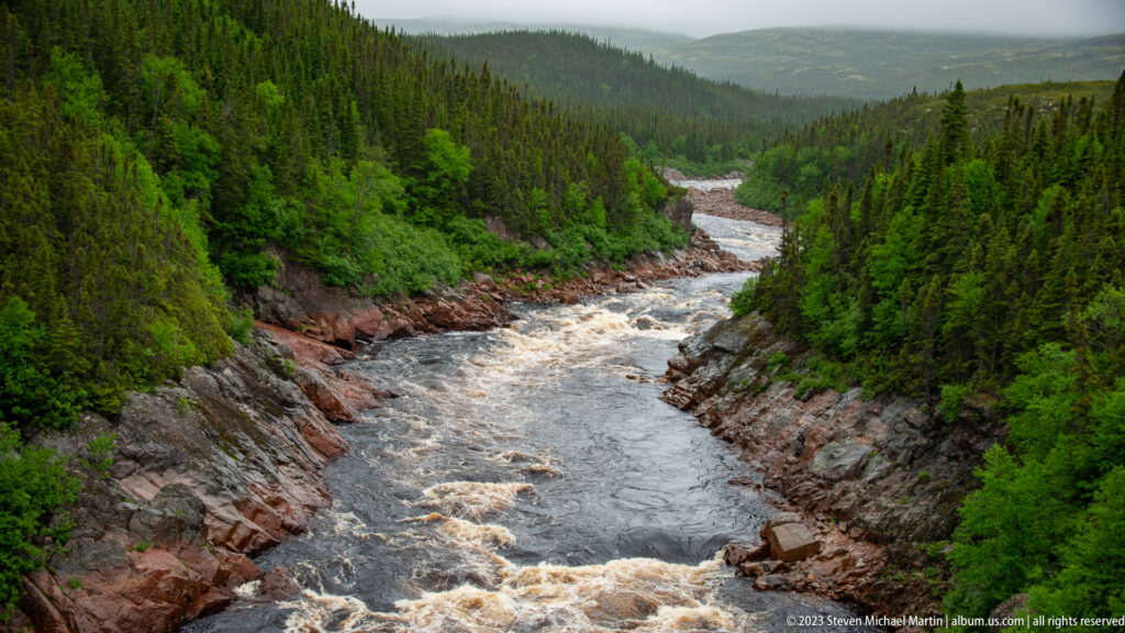 Rapids running through a reddish rock gorge surrounded by dense spruce forest and misty hills.