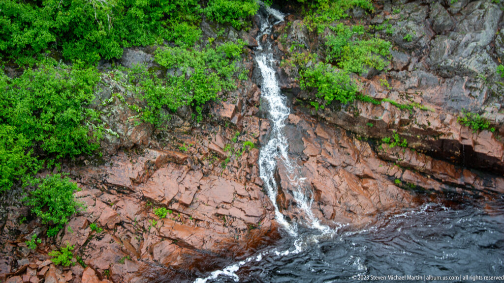 Narrow waterfall cascading down reddish fractured rock into dark river water amid bright green shrubs.