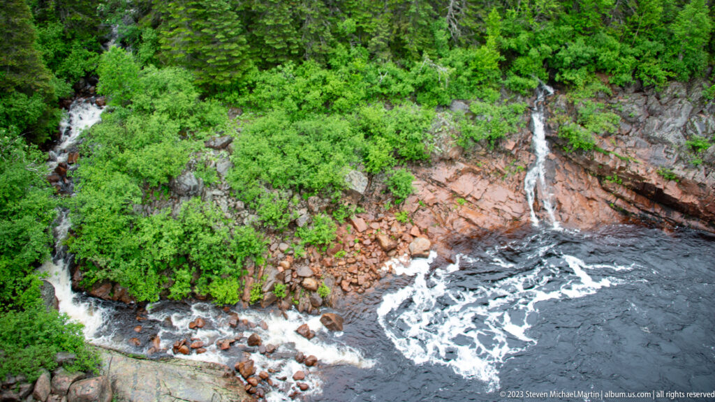 Two small cascades descend through green vegetation and reddish rocks into a dark river edged with white foam.