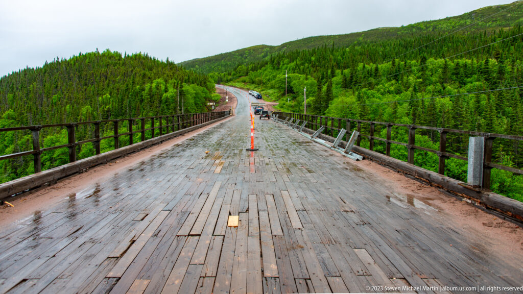 Wet wooden bridge deck with metal railings leading toward a forested road curve and low green hills.