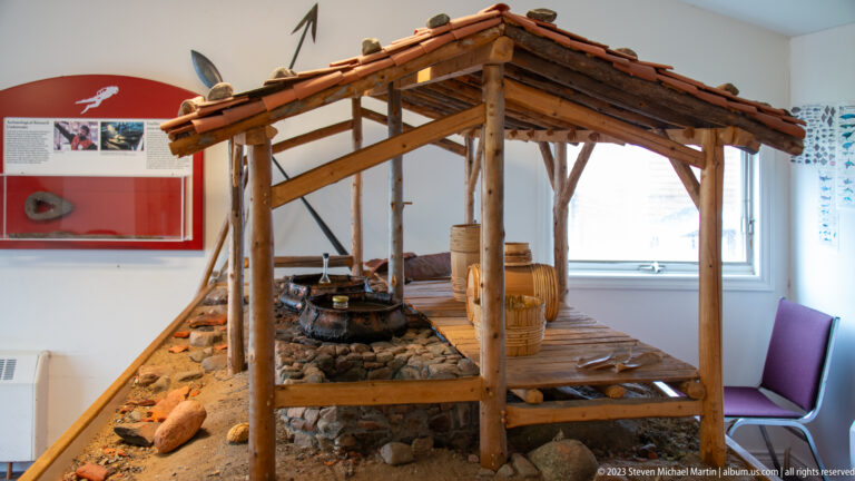 Indoor museum reconstruction of a timber shelter with pitched roof, wooden barrels, plank floor, and stone-ringed hearth.