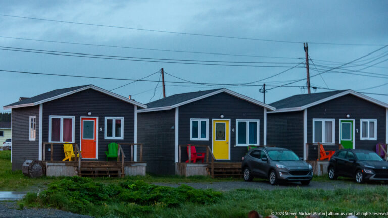 Three dark gabled cottages with red, yellow, and green doors, colorful porch chairs, parked cars, and utility lines.