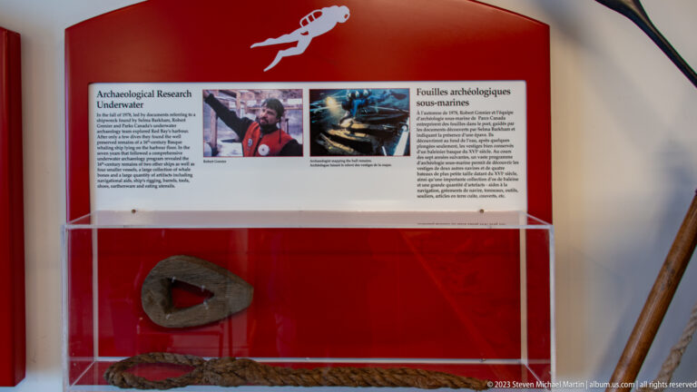 Bilingual Red Bay museum display about underwater archaeology with diver photographs and artifacts in a glass case.