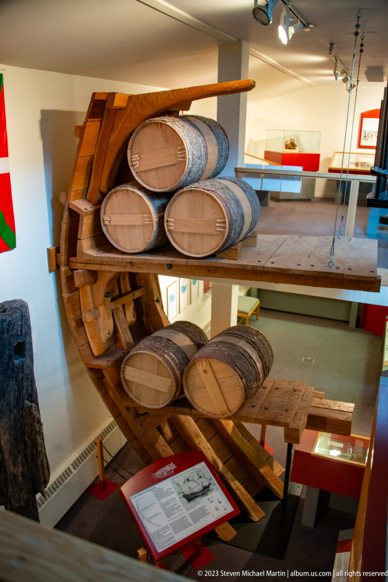 Cutaway timber museum reconstruction with stacked wooden barrels, heavy framing, plank floors, and a stone work area.