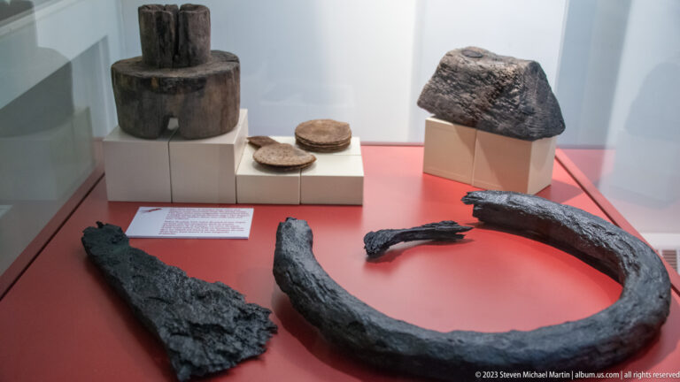 Red museum case containing corroded iron and wooden archaeological artifacts displayed on white mounts.