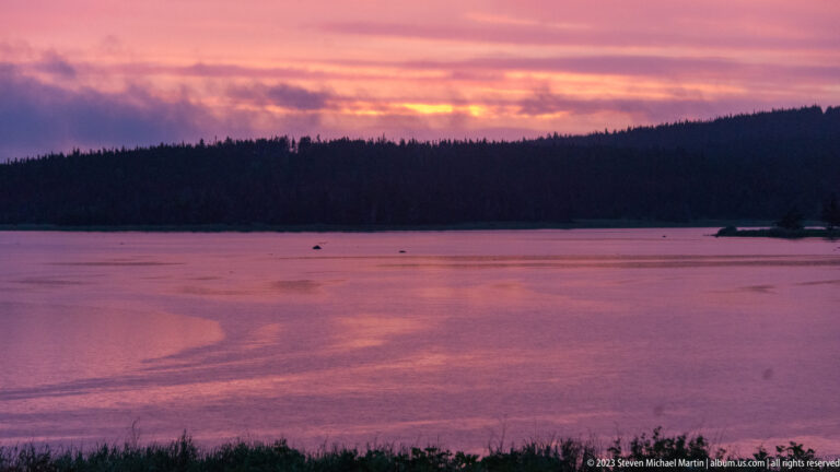 Calm water tinted pink and purple at dusk, with a dark forested shoreline beneath layered clouds.
